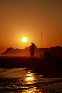 Silhouette man standing at beach against clear orange sky during sunset