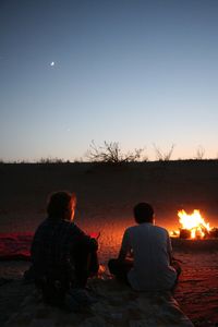 Rear view of men sitting by bonfire against sky at sunset