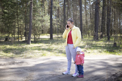 Full length of mother talking on phone standing with daughter against trees