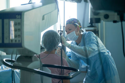 Attentive female surgeon in sterile uniform examining eye of anonymous patient against refractometer in hospital