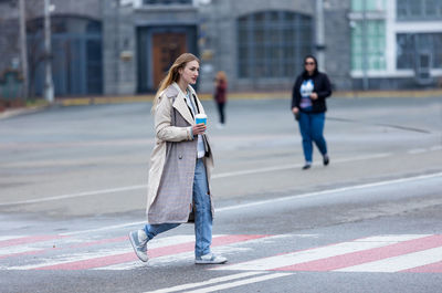 Shot of a pretty young woman in coat crossing the street holding coffee cup.