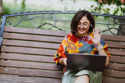 An adult asian woman in a park on bench communicates via video call on a laptop