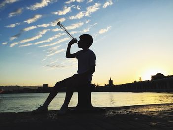 Boy with drink while sitting on bollard at pier during sunset