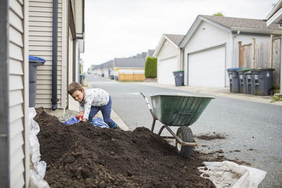 Young boy scooping pile of soil into wheelbarrow in back alley