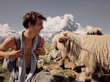 Happy young man sitting by sheep against sky