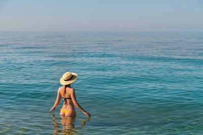 Back view of a slender caucasian young woman standing waist-deep in calm transparent sea water in a