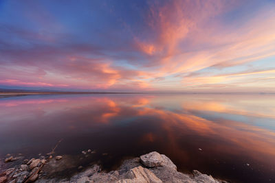 Rocks by lake with clouds reflection against sky during sunset