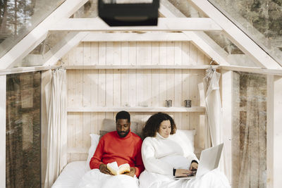 Woman working on laptop while boyfriend reading book in hotel