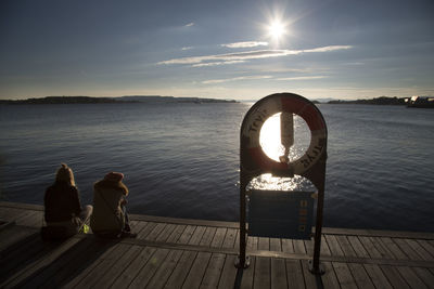 Rear view of women sitting on pier over lake against sky during sunset