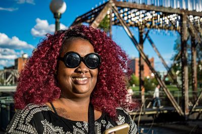 Portrait of woman with curly maroon hair while wearing sunglasses against bridge