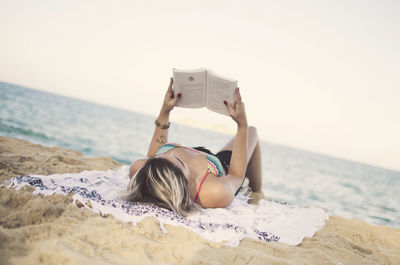 Woman reading book while relaxing at beach against sky