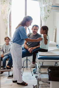 Pediatrician checking girl with stethoscope while sitting with family in medical examination room