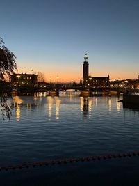 Illuminated buildings by river against sky at sunset