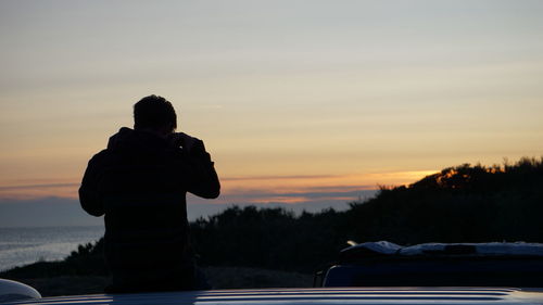 Rear view of silhouette man sitting on car against sky during sunset