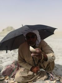 Man holding seashell while sitting with umbrella at beach