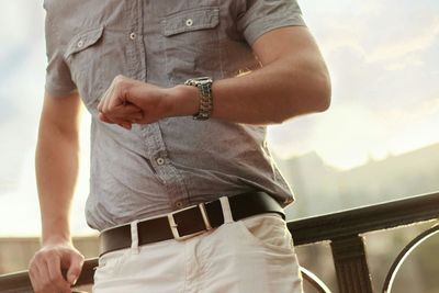 Midsection of man checking time while standing against railing during sunset