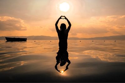 Silhouette woman forming heart shape in sea against sky during sunset