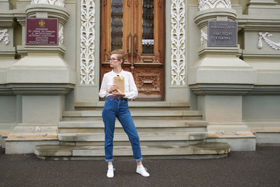 Full length of young woman holding book standing against building