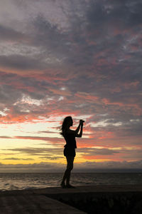 Silhouette woman photographing at beach against sky during sunset