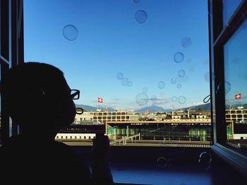 Close-up of boy with bubbles looking through window against blue sky