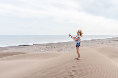 Vertical photo of a woman with sunglasses using a portable tripod to take a selfie in a dune