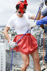 Woman wearing hero costume being prepared by a man to descend a tall rappel building. 