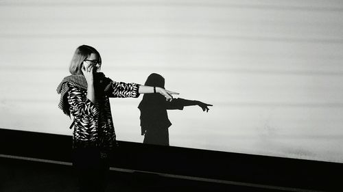 Woman pointing while talking on mobile phone against wall