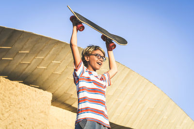 Low angle view of smiling boy holding skateboard standing against sky