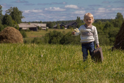The boy with the suitcase is smiling. he's coming into the field. around the haystack, a village