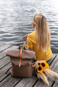 A young woman sits on a pier by the lake next to a traveler's backpack and a bouquet of sunflowers.