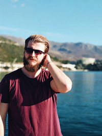 Portrait of bearded young man wearing sunglasses against lake and sky