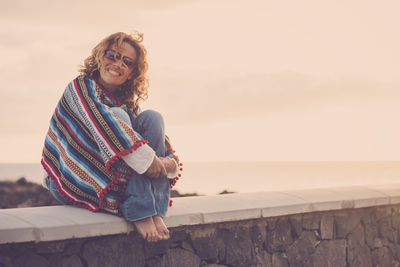 Smiling woman sitting on retaining wall by sea against sky