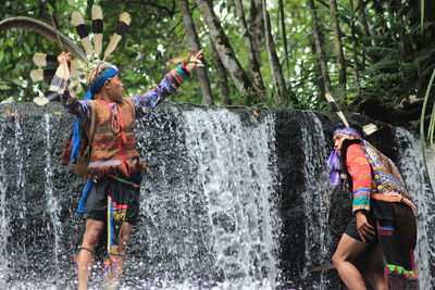 People wearing traditional clothing standing in waterfall against trees at forest