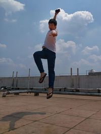 Full length portrait of young man gesturing while holding mobile phone on building terrace against sky