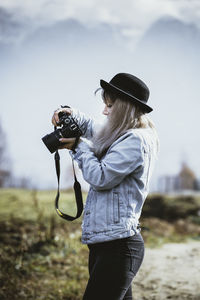 Side view of young woman photographing through camera against sky