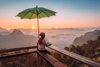 Rear view of man standing on railing against sky during sunset