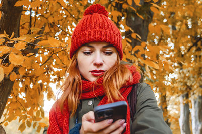 Stylish girl with mobile phone in autumn park. orange knitted hat and scarf, bright lipstick.