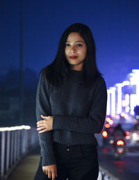 Portrait of young woman standing against illuminated city at night