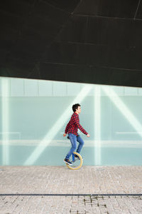 Full body side view of active young male in checkered shirt and jeans performing trick on unicycle near mirrored glass wall of contemporary building