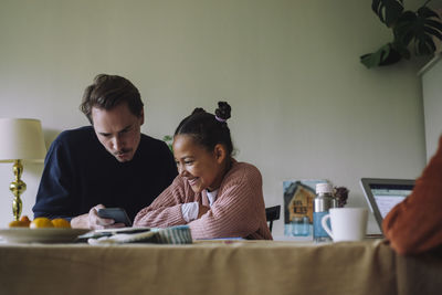 Gay man sharing smart phone with happy daughter sitting at dining table in home