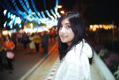 Portrait of beautiful smiling woman standing on sidewalk in illuminated city at night