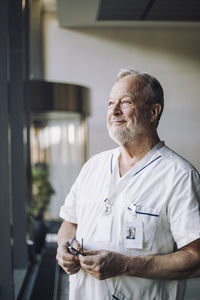 Thoughtful senior male doctor in scrubs holding eyeglasses while standing at hospital