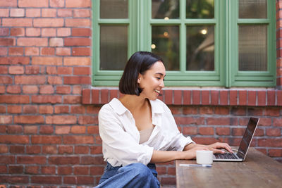 Side view of young woman using mobile phone while standing against brick wall