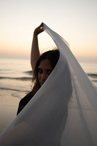 Portrait of young woman standing at beach against sky during sunset