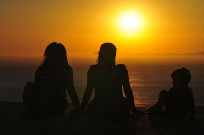 Silhouette people relaxing at beach against sky during sunset