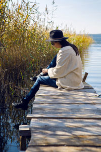 Woman in a black hat and sweater sits on the old pier to the bushes by the lake