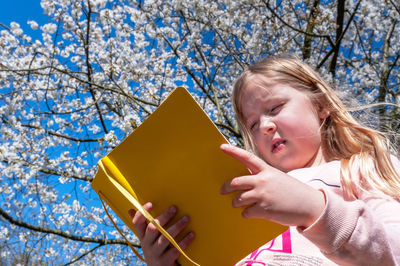 Little girl sitting against blooming spring tree and reading from yellow book.