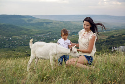 Woman with a child in a field on the mountain sitting next to a small goat in the summer