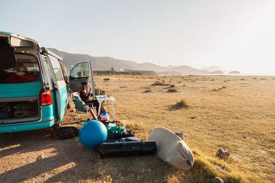 Woman sitting on chair by camper van against sky