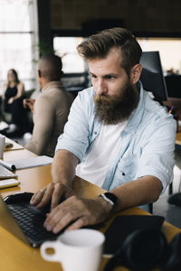 Bearded male computer programmer working on laptop at desk in office